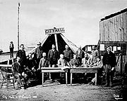 Los hombres de pie y sentados alrededor de dos mesas, frente a la cámara. Una gran carpa detrás de ellos tiene un cartel de madera que lee