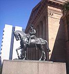 Estatua ecuestre de metal en gris oscuro de George V vestido de militar uniforme sobre un pedestal de granito rojo fuera de un edificio clásico de la piedra arenisca roja