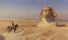 Persona en un caballo mira hacia una gigantesca estatua de una cabeza en el desierto, con un cielo azul