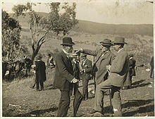 Tres hombres de mediana edad con barba corta en trajes formales y sombreros de pie en el campo montañoso abierto con un solo árbol cercano