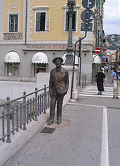 Estatua de bronce de Joyce pie en una acera, junto a una barandilla. Detrás de la estatua es una escena de la calle con los peatones y tiendas.
