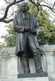 Una escultura de metal de bronce de un hombre del siglo XIX con un largo, chaqueta o abrigo, pantalones, chaleco, con herramientas de dibujante en sus manos
