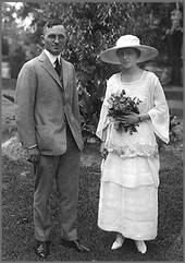 Fotografía de boda de Truman en traje gris y su mujer en el sombrero con vestido blanco tomarse de las flores