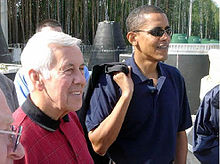 Hombre de pelo gris y Obama destacan, vistiendo camisas de polo ocasionales. Obama lleva gafas de sol y tiene algo colgando de su hombro derecho.