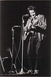 Dylan con su guitarra en el escenario, riendo y mirando hacia abajo.