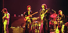 Dylan junto a tres músicos de la banda en el escenario. Dylan es el tercero desde la izquierda, que llevaba un saco negro y pantalones. Está cantando y tocando una guitarra eléctrica.