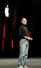 Retrato de cuerpo entero del hombre de unos cincuenta con pantalones vaqueros y una camisa de cuello alto negro, de pie delante de una cortina oscura con un logotipo blanco de Apple